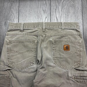 Puede incluir: Primer plano de la parte trasera de unos pantalones cargo verde oliva. Los pantalones tienen dos bolsillos traseros y un parche de cuero marr&oacute;n con un logotipo. Los pantalones tienen trabillas y cierre de bot&oacute;n. La tela parece ser una mezcla de algod&oacute;n duradera.