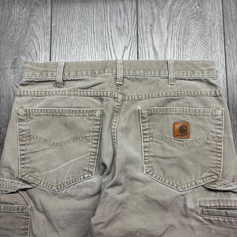 Puede incluir: Primer plano de la parte trasera de unos pantalones cargo verde oliva. Los pantalones tienen dos bolsillos traseros y un parche de cuero marr&oacute;n con un logotipo. Los pantalones tienen trabillas y cierre de bot&oacute;n. La tela parece ser una mezcla de algod&oacute;n duradera.