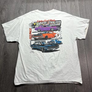 May include: A white short-sleeved t-shirt featuring a graphic print of four classic Dodge muscle cars in purple, orange, and blue. The text reads Dodge Scat Pack Boulevard, 440 Six Pack, and Hemi. The design includes illustrations of smoke and speed.