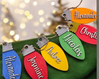 May include: A garland of wooden Christmas light bulb ornaments in blue, red, yellow, green, and orange. Each bulb features a grey base and a white script name attached to the front. The ornaments are connected by a thin brown twine string.