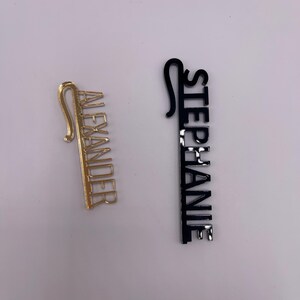 May include: Two name tags, one gold and one black. The gold tag reads "ALEXANDER" in a vertical font. The black tag reads "STEPHANIE" in a vertical font. Both tags have a hook at the top.