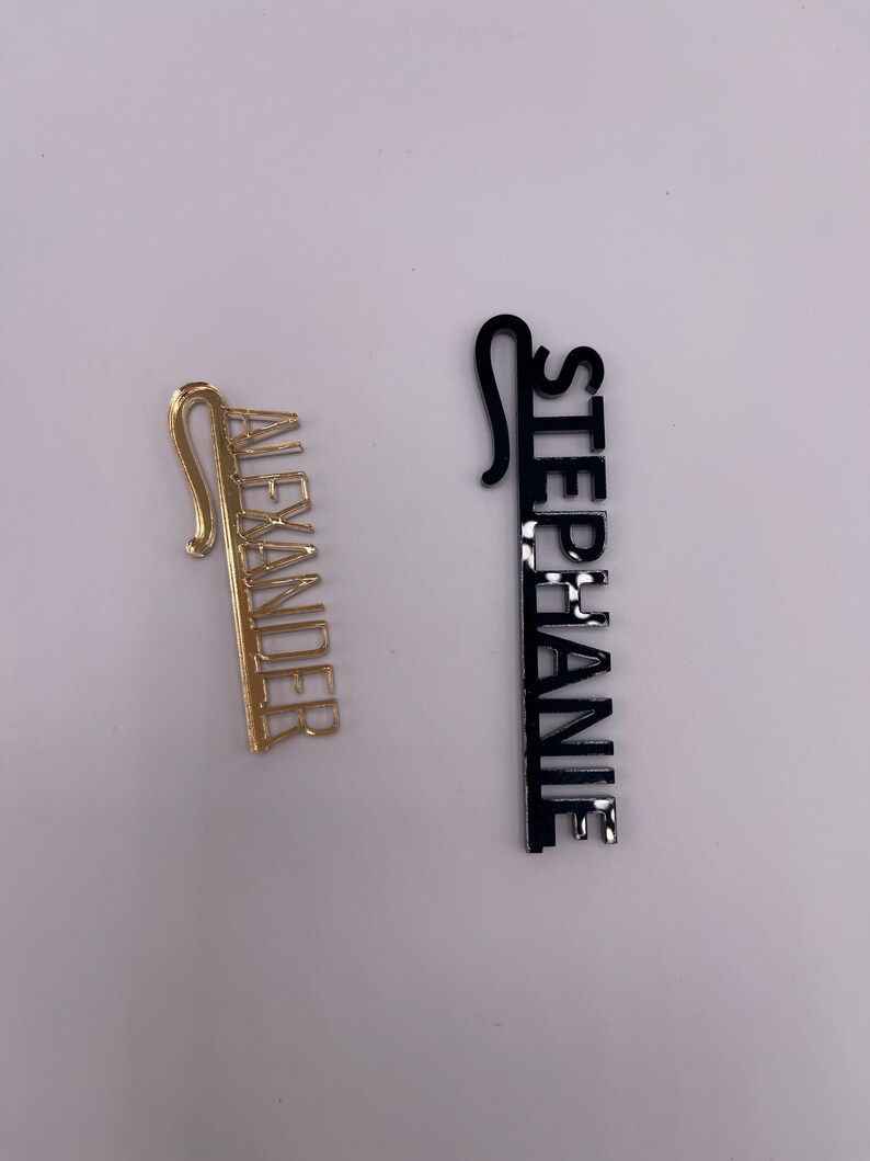 May include: Two name tags, one gold and one black. The gold tag reads "ALEXANDER" in a vertical font. The black tag reads "STEPHANIE" in a vertical font. Both tags have a hook at the top.