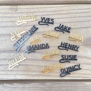 May include: A collection of personalized name tags in various colors, including gold, black, and silver. Each tag features a different name in a stylish font, with a curved design element. The tags are arranged on a wooden surface.