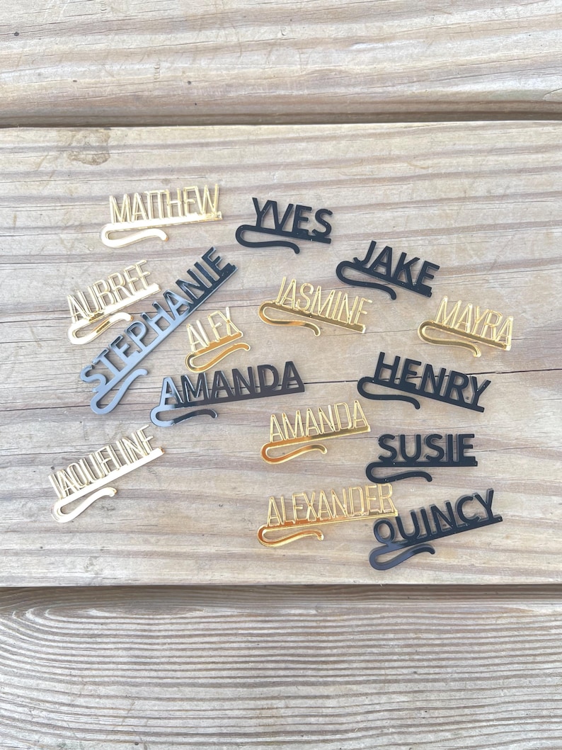 May include: A collection of personalized name tags in various colors, including gold, black, and silver. Each tag features a different name in a stylish font, with a curved design element. The tags are arranged on a wooden surface.