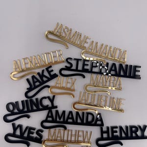 May include: A collection of name tags in gold and black. The tags feature various names in a stylish font, with a curved bar for attachment. The names include "Jasmine", "Amanda", "Alexander", "Jake", "Quincy", and others.