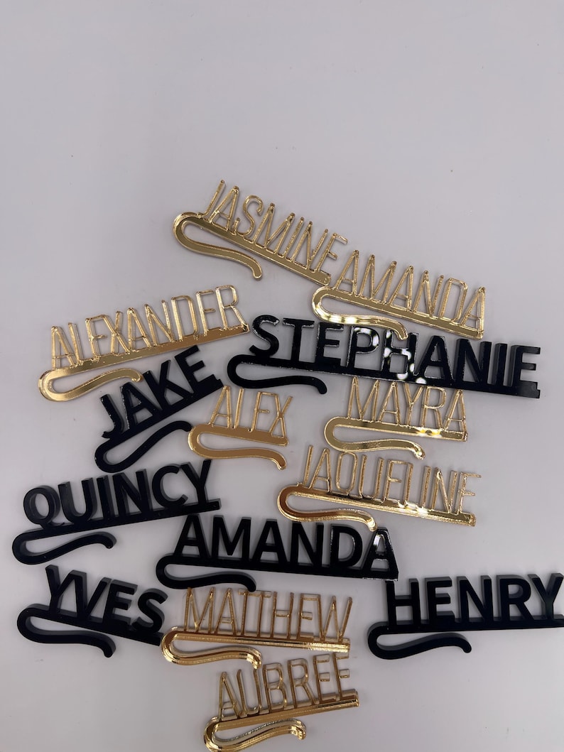 May include: A collection of name tags in gold and black. The tags feature various names in a stylish font, with a curved bar for attachment. The names include "Jasmine", "Amanda", "Alexander", "Jake", "Quincy", and others.