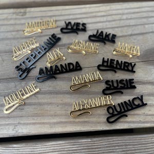May include: A collection of personalized name pins in gold and black. The pins feature various names in a cursive font, including "Matthew," "Aubree," and "Amanda." The pins are arranged on a wooden surface.