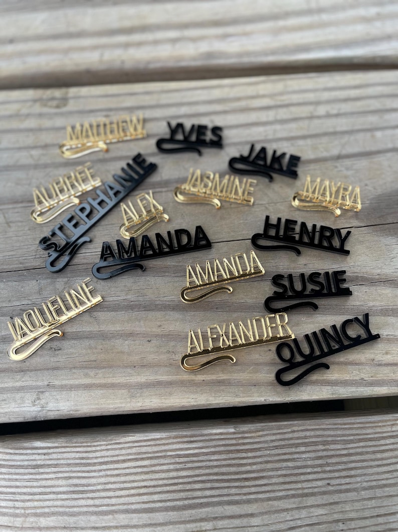 May include: A collection of personalized name pins in gold and black. The pins feature various names in a cursive font, including "Matthew," "Aubree," and "Amanda." The pins are arranged on a wooden surface.