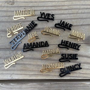 May include: A collection of personalized name tags in gold and black. The names are in a stylish font with a curved clip design. Names include Matthew, Yves, Jake, and others, displayed on a wooden surface.