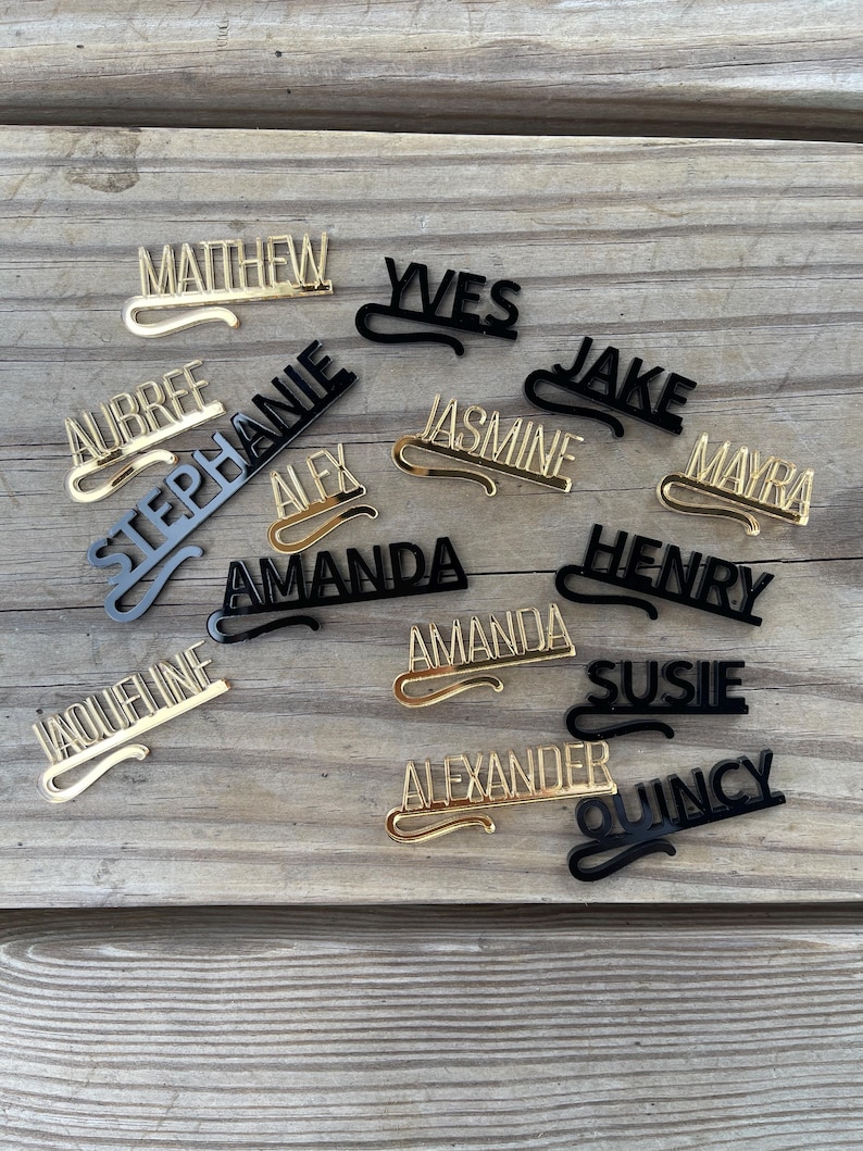 May include: A collection of personalized name tags in gold and black. The names are in a stylish font with a curved clip design. Names include Matthew, Yves, Jake, and others, displayed on a wooden surface.