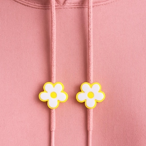 May include: Close-up of a pink hoodie with a drawstring and two white and yellow flower-shaped charms. The hoodie's hood is visible at the top, and the drawstring is threaded through the hood.