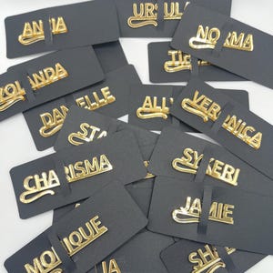 May include: A collection of gold-colored name pins, each with a different name, displayed on black rectangular cards. The names are in a stylish, cursive font. The pins are scattered, creating a visually appealing arrangement.