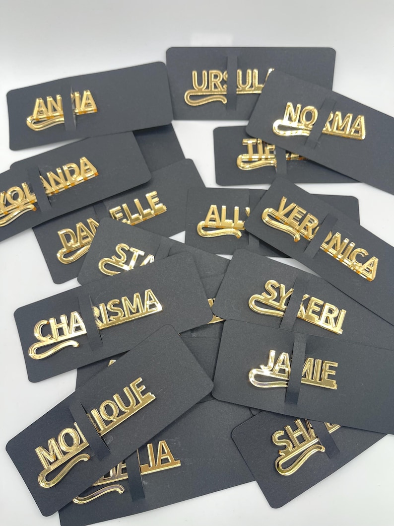 May include: A collection of gold-colored name pins, each with a different name, displayed on black rectangular cards. The names are in a stylish, cursive font. The pins are scattered, creating a visually appealing arrangement.
