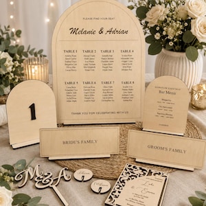 May include: A set of light wood wedding stationery including an arched seating chart labeled Melanie & Adrian, a table number one sign, a bar menu, two rectangular family signs, a cake topper with M&A, two small circular name tags, and an invitation.