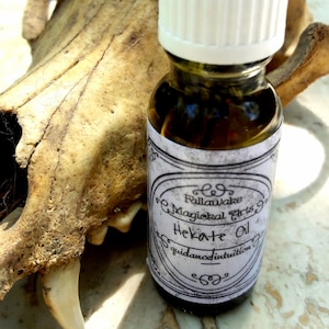 Könnte beinhalten: Eine kleine Glasflasche mit weißem Verschluss und einem Etikett mit der Aufschrift "Fullawake Magickal Arts Hekate Oil guidance/intuition". Die Flasche steht auf einer hellbraunen Oberfläche.