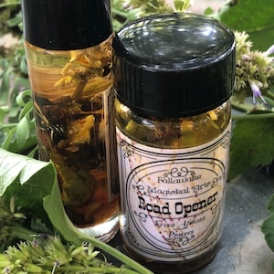 May include: Two glass bottles filled with a golden liquid and botanical elements. One is a roll-on bottle, the other a jar with a black cap. The jar has a label that reads "Road Opener" and "Magickal Arts". Green foliage surrounds the bottles.
