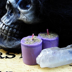 May include: A spooky still life featuring a skull, two purple candles topped with herbs, and a clear quartz crystal. The items are arranged on a Ouija board, creating a mystical and gothic aesthetic.