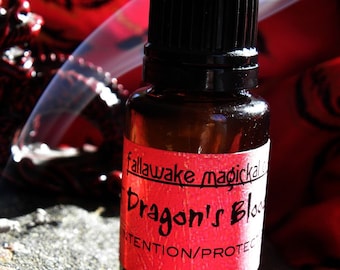 May include: A small, brown glass bottle with a black cap. A red label reads "fallawake magickals Dragon's Blood INTENTION/PROTECT" in black script. The bottle is on a textured surface with a red fabric background.