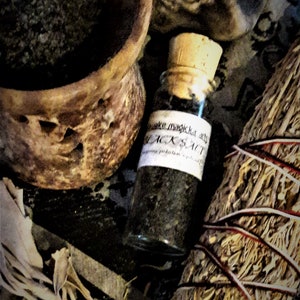 Könnte beinhalten: Eine kleine Glasflasche mit einem Korkverschluss, gefüllt mit schwarzem Salz. Die Flasche trägt ein Etikett mit der Aufschrift "Make magick with BLACK SALT". Die Flasche steht auf einer Holzoberfläche mit einem Salbeistab und einer Tonschale.