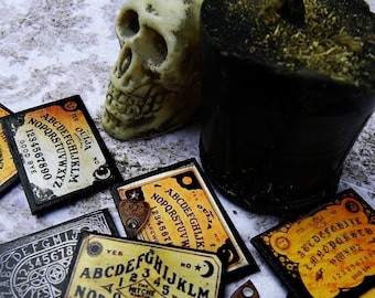 May include: A collection of vintage Ouija boards with a black candle and a white skull. The boards are yellow and black with the letters of the alphabet, numbers, and the words "yes", "no", and "goodbye".