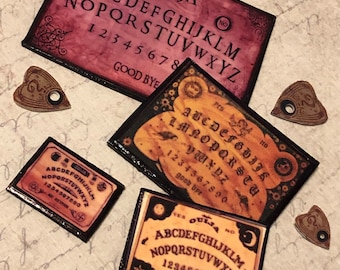 May include: Four miniature Ouija boards with planchette pointers. The boards are in various colors and designs, including red, black, and gold. The boards feature the letters of the alphabet, numbers, and the words "Yes", "No", "Goodbye", and "Farewell".