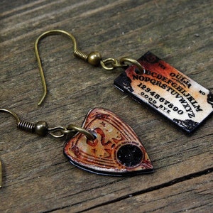 May include: A pair of earrings featuring a heart-shaped pendant with a Ouija board design and a rectangular pendant with a traditional Ouija board design. The pendants are made of metal and have a distressed, vintage look. The earrings are attached to gold-toned hooks.