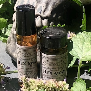 May include: Two small glass bottles with black caps, labeled "HEX AWAY" with decorative text. The bottles are filled with a light amber liquid. The background includes a skull and greenery, suggesting a mystical or apothecary theme.