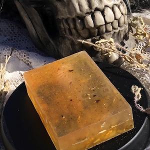 May include: A translucent, amber-coloured soap bar with flecks of dried herbs and flowers. The soap is sitting on a black circular surface.