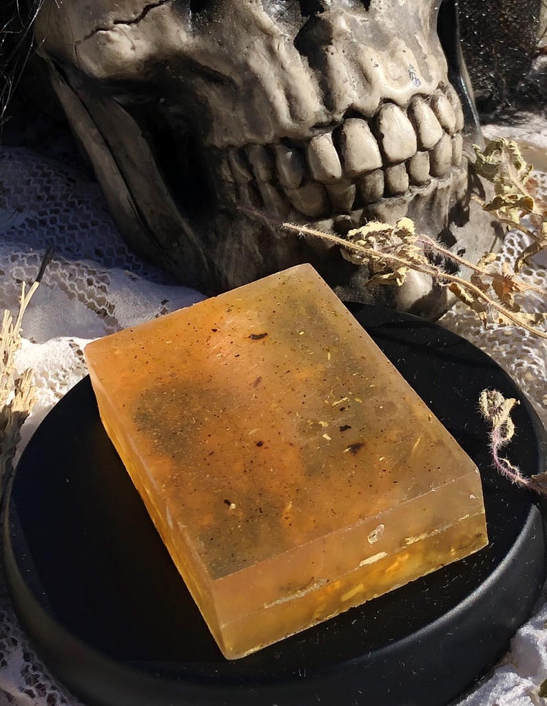 May include: A translucent, amber-coloured soap bar with flecks of dried herbs and flowers. The soap is sitting on a black circular surface.