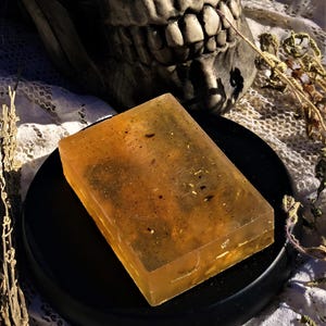 Może przedstawiać: Przezroczysty, bursztynowy kawałek mydła z drobinkami ziół i kwiatów w środku. Mydło leży na czarnym, okrągłym talerzu.