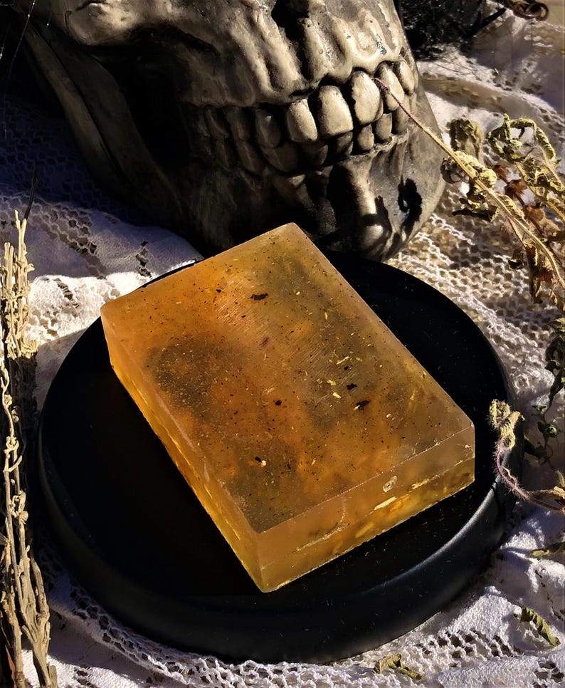 May include: A translucent amber-coloured bar of soap with flecks of herbs and flowers inside. The soap is sitting on a black circular dish.