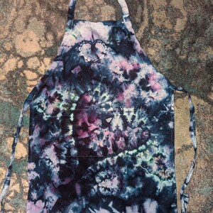 May include: A tie-dye apron with a dark blue, purple, and white color scheme. The apron has a single pocket on the front and adjustable straps.
