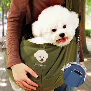 Pode incluir: Uma bolsa de transporte para animais de estimação verde-oliva com um cão Bichon Frise branco dentro. A bolsa tem um pequeno bordado do rosto do cão e o nome "Snowy". Um bolso para telefone embutido é visível.