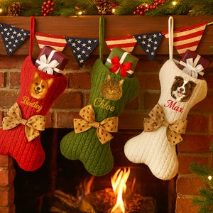 Op de afbeelding: Drie gepersonaliseerde kerstsokken in rood, groen en wit, elk met het gezicht en de naam van een huisdier geborduurd. Elke sok is versierd met een strik en gevuld met een ingepakt cadeau. Feestelijke vakantiedecoratie.
