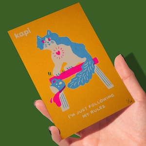 May include: A mustard yellow art print featuring a whimsical cat illustration with blue and white fur, a pink heart, and a pink and blue object. The text "kapi" is at the top left, and "I'M JUST FOLLOWING MY RULES" is at the bottom.