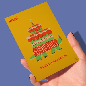 May include: A yellow birthday card with a turtle-shaped cake illustration. The card features the text "kapi" in red at the top left and "SHELL-EBRATEION" at the bottom. The turtle has a party hat.
