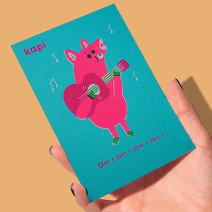 May include: A teal card featuring a pink pig playing a pink guitar. The pig has green feet and is surrounded by musical notes. The word "kapi" is in the upper left corner, and the phrase "OH I OH I OH I OH !" is at the bottom.