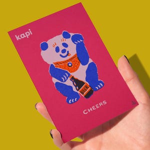 May include: A pink card featuring a cartoon panda holding a beer bottle. The panda is blue and white with an orange neckerchief. The word "kapi" is in the upper left corner, and "CHEERS" is at the bottom.