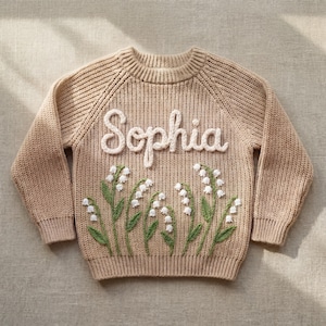 Puede incluir: Un su&eacute;ter de punto beige con el nombre "Sophia" en escritura blanca en el pecho. Debajo del nombre hay un dise&ntilde;o bordado de flores de lirio de los valles con tallos verdes y flores blancas. El su&eacute;ter tiene mangas largas.