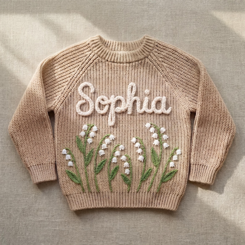 Puede incluir: Un su&eacute;ter de punto beige con el nombre "Sophia" en escritura blanca en el pecho. Debajo del nombre hay un dise&ntilde;o bordado de flores de lirio de los valles con tallos verdes y flores blancas. El su&eacute;ter tiene mangas largas.