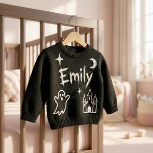 May include: Black knit sweater with white embroidered Halloween-themed designs, including a ghost, castle, stars, and moon. The name "Emily" is embroidered in white. The sweater is hanging on a wooden hanger.