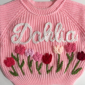 A pink knit sweater with the name "Dahlia" embroidered in a cursive font. The sweater features embroidered tulips in shades of pink and red with green stems and leaves.