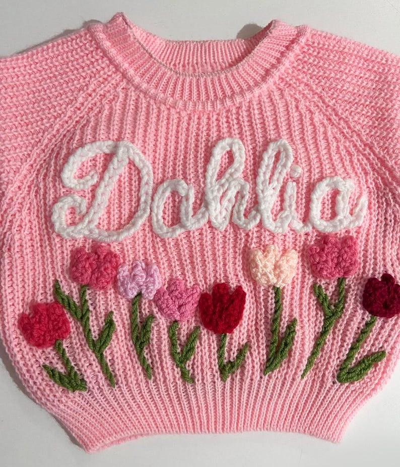 A pink knit sweater with the name "Dahlia" embroidered in a cursive font. The sweater features embroidered tulips in shades of pink and red with green stems and leaves.