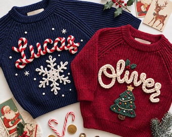 May include: A navy blue knit jumper with the name Lucas in red and white candy cane yarn and a white snowflake, and a red knit jumper with the name Oliver in white yarn and a green Christmas tree with colourful ornaments and a gold star.