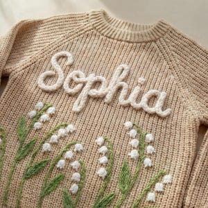 Puede incluir: Un su&eacute;ter de punto beige con el nombre "Sophia" en letras blancas y cursivas. El su&eacute;ter presenta un dise&ntilde;o bordado de flores blancas de lirios de los valles con tallos y hojas verdes.