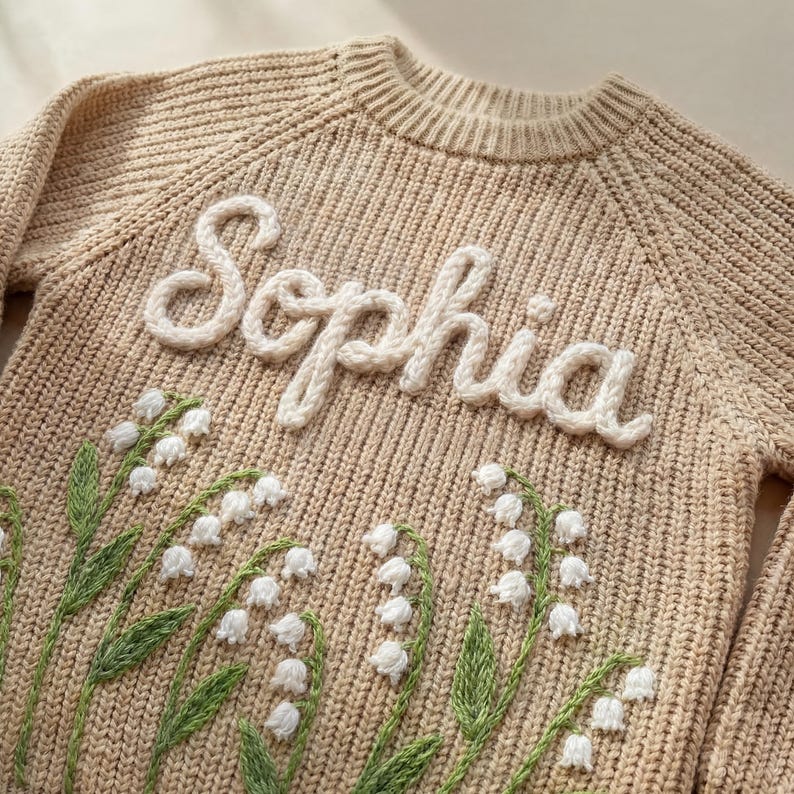 Puede incluir: Un su&eacute;ter de punto beige con el nombre "Sophia" en letras blancas y cursivas. El su&eacute;ter presenta un dise&ntilde;o bordado de flores blancas de lirios de los valles con tallos y hojas verdes.