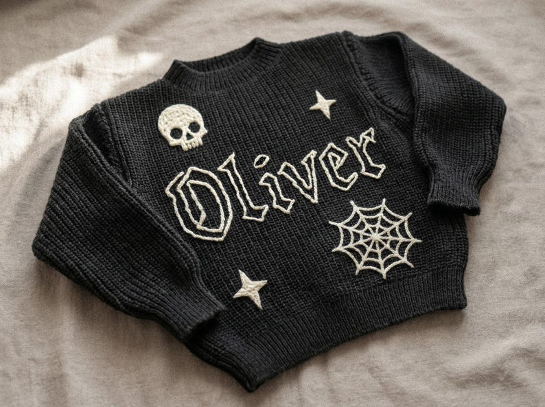 May include: Black knit sweater with the name "Oliver" in white embroidered letters. The sweater features a skull, stars, and a spiderweb design. A cozy, Halloween-themed garment.