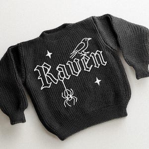 May include: Black knit sweater with the word "Raven" in white gothic lettering, a white raven, a spider, and two white stars. The sweater has long sleeves and a crew neck.