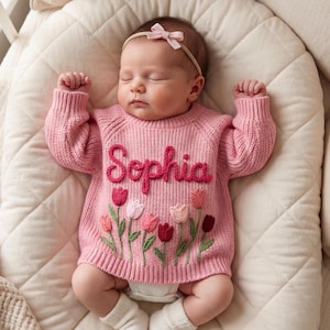 Puede incluir: Un beb&eacute; con un su&eacute;ter de punto rosa con el nombre "Sophia" bordado en rosa oscuro. El su&eacute;ter presenta un dise&ntilde;o floral con tulipanes en tonos rosa, rojo y blanco. El beb&eacute; tambi&eacute;n lleva una diadema con lazo rosa y calcetines blancos.