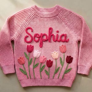 A pink knit sweater with the name "Sophia" embroidered in a cursive font. The sweater features embroidered tulips in shades of pink and red with green stems and leaves.